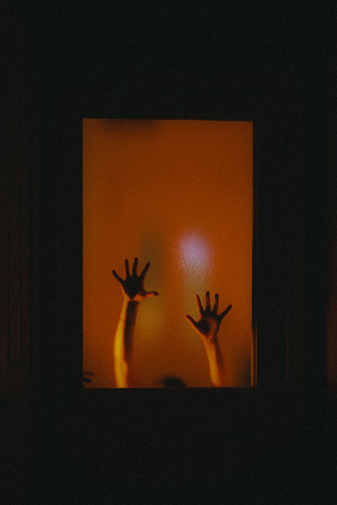 Mysterious silhouette of raised hands behind frosted glass with a warm glow.